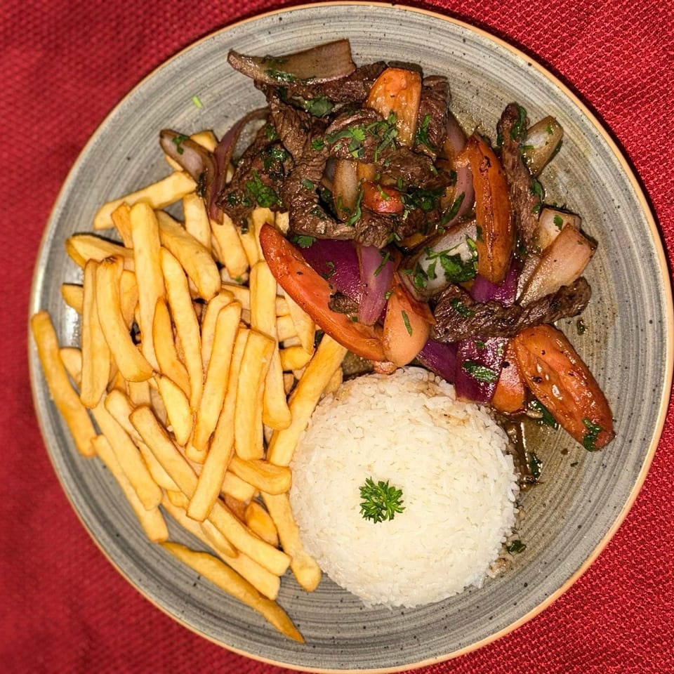 Lomo Saltado with french fries and white rice on a gray plate, against a red background.